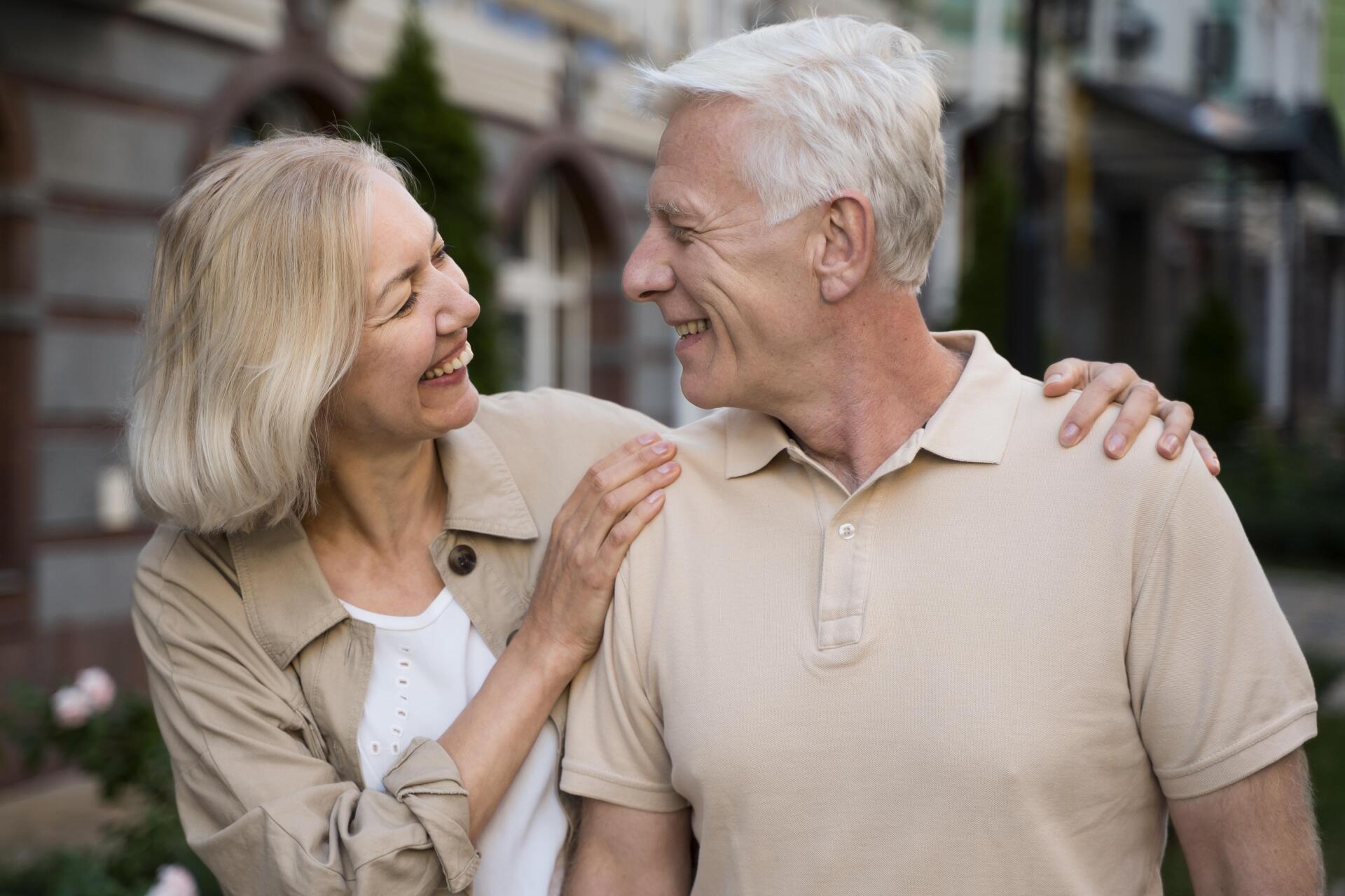 Smiling couple outdoors, woman's arm around man's shoulders, looking at each other.