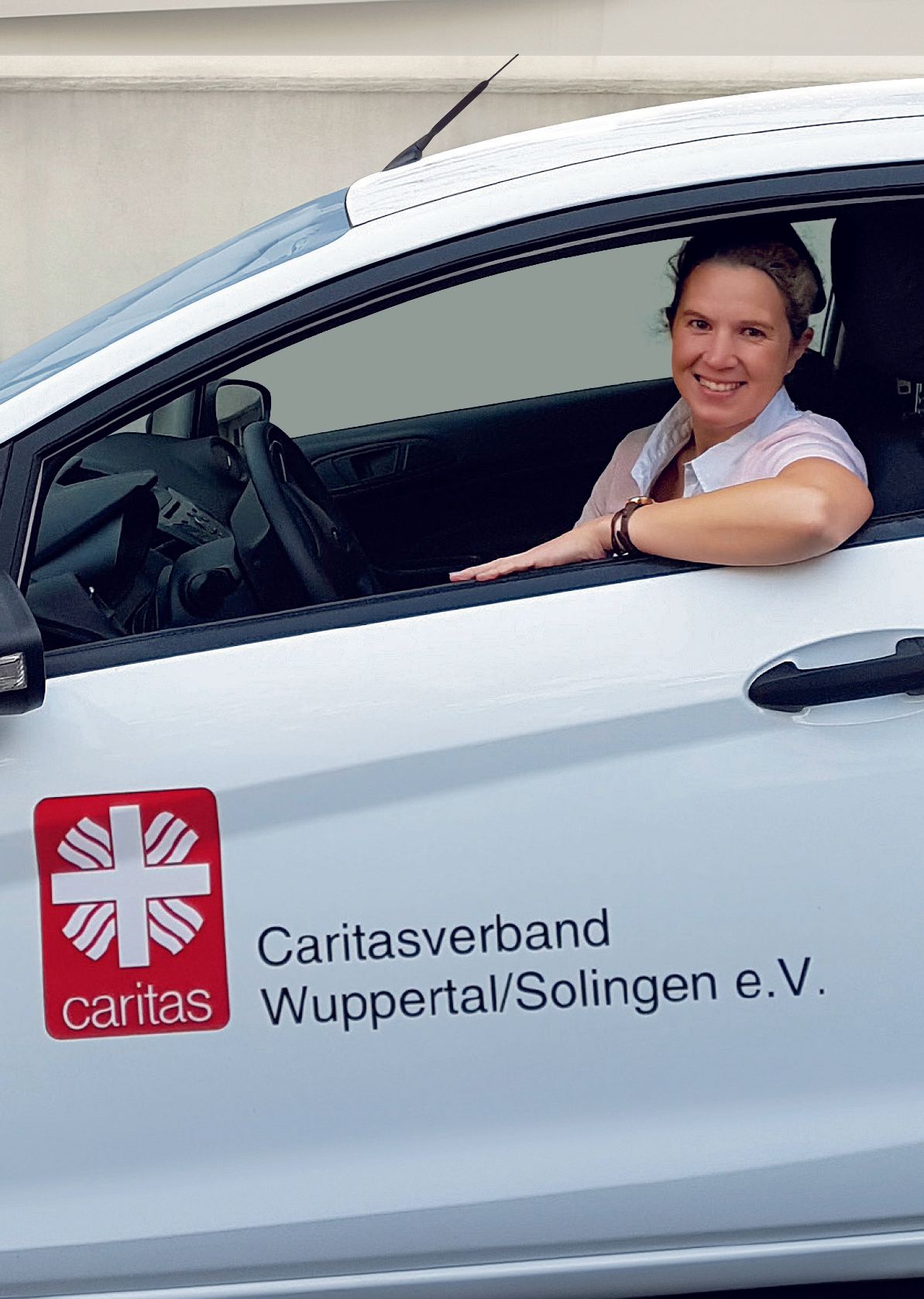 Lächelnde Frau in einem weißen Auto mit Caritas-Logo auf der Tür.
