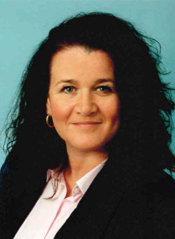 Frau mit dunklem lockigem Haar, rosa Hemd und schwarzem Blazer lächelt.