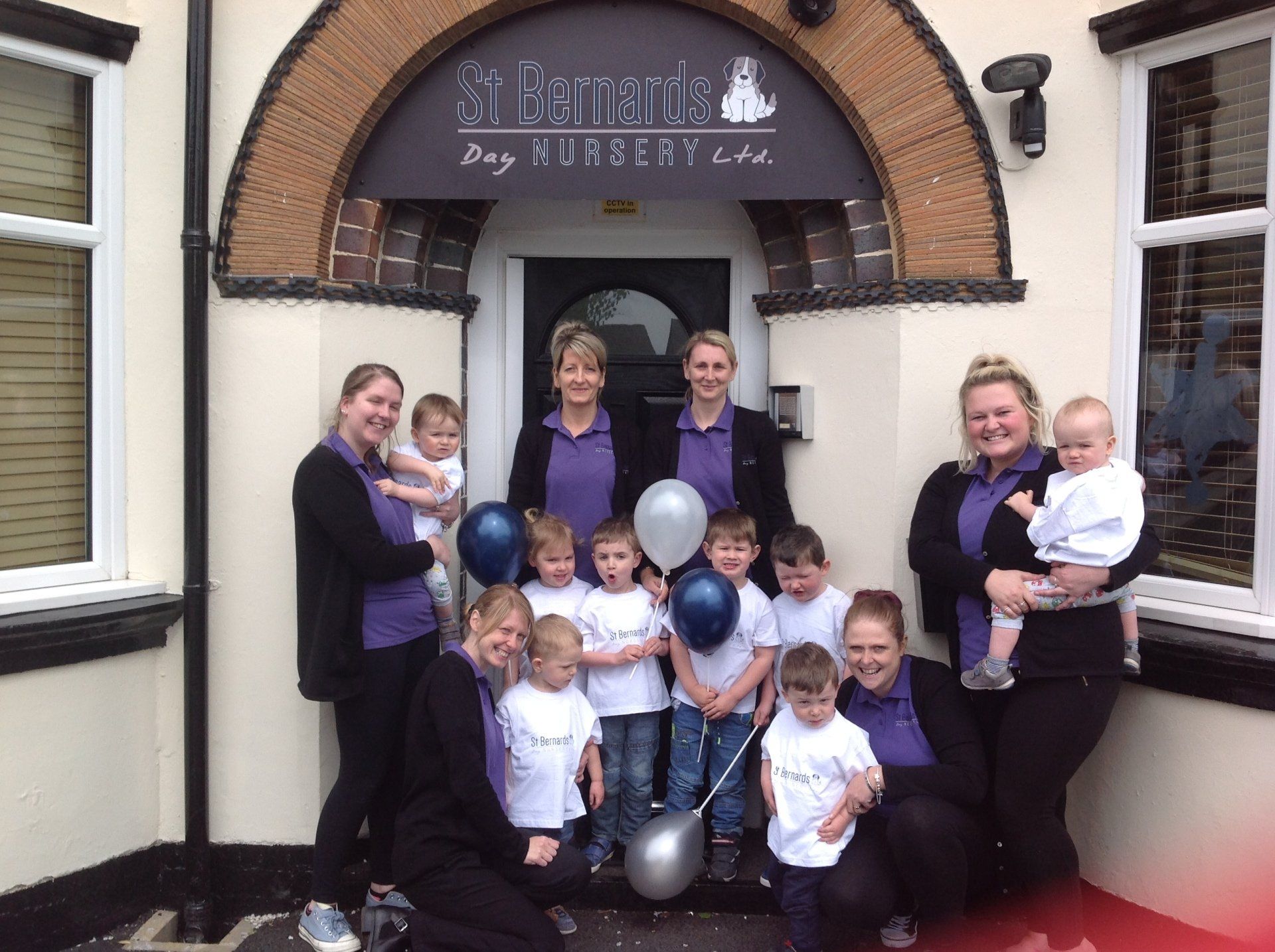 Group posing outside a bakery, with balloons and children holding babies in front of the doorway