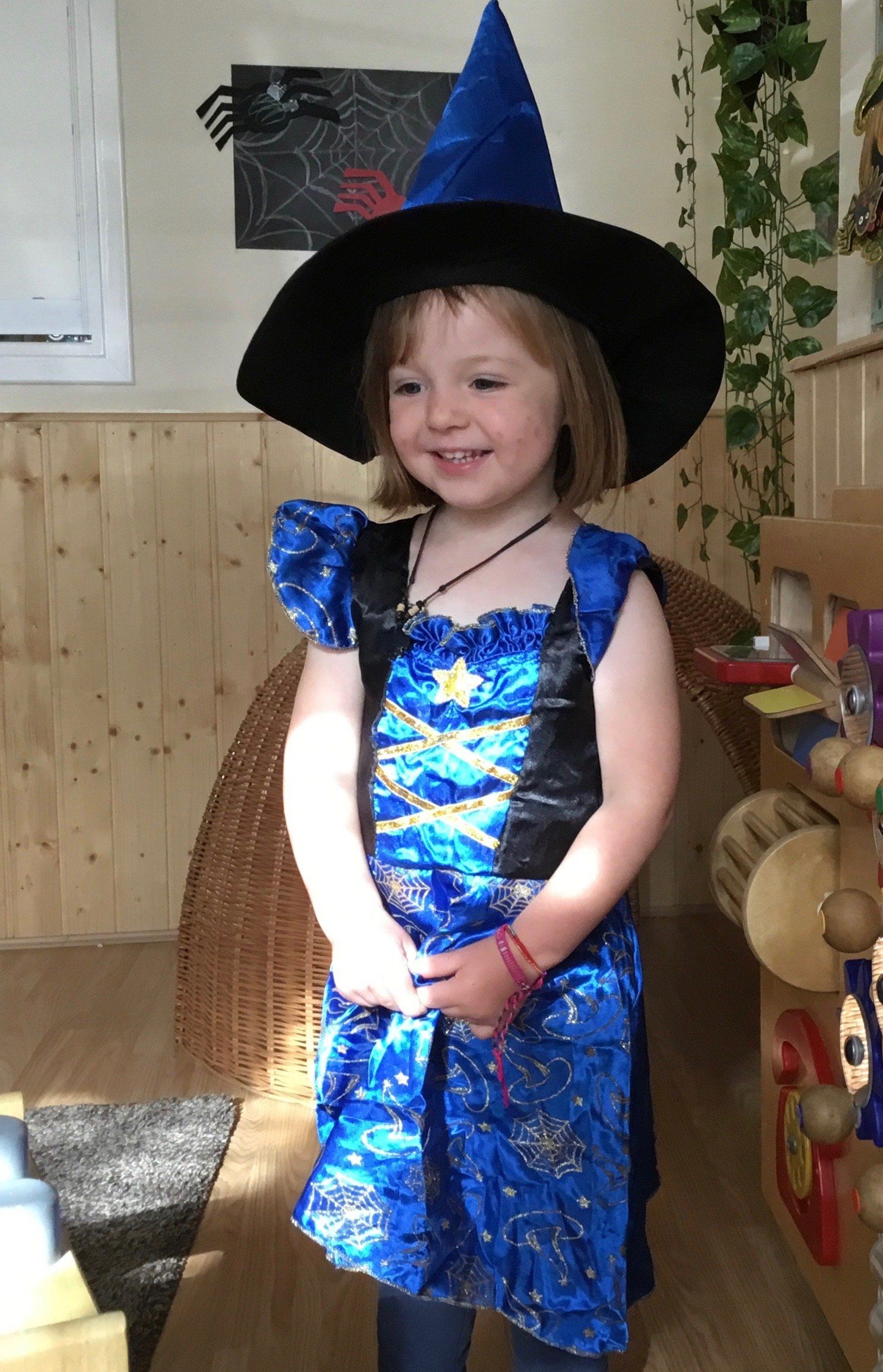 Child in a blue witch costume and black hat, smiling indoors.
