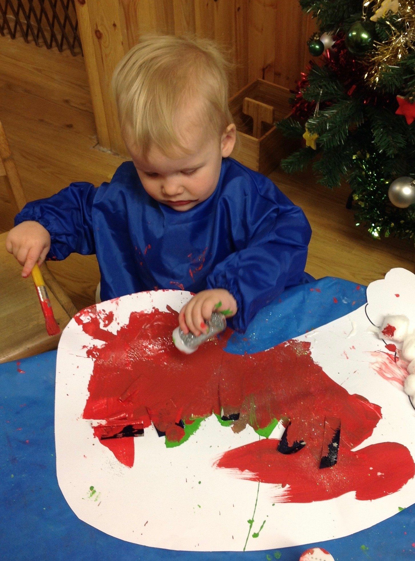 Child painting a large red shape on white paper with a brush, indoors near a decorated tree.