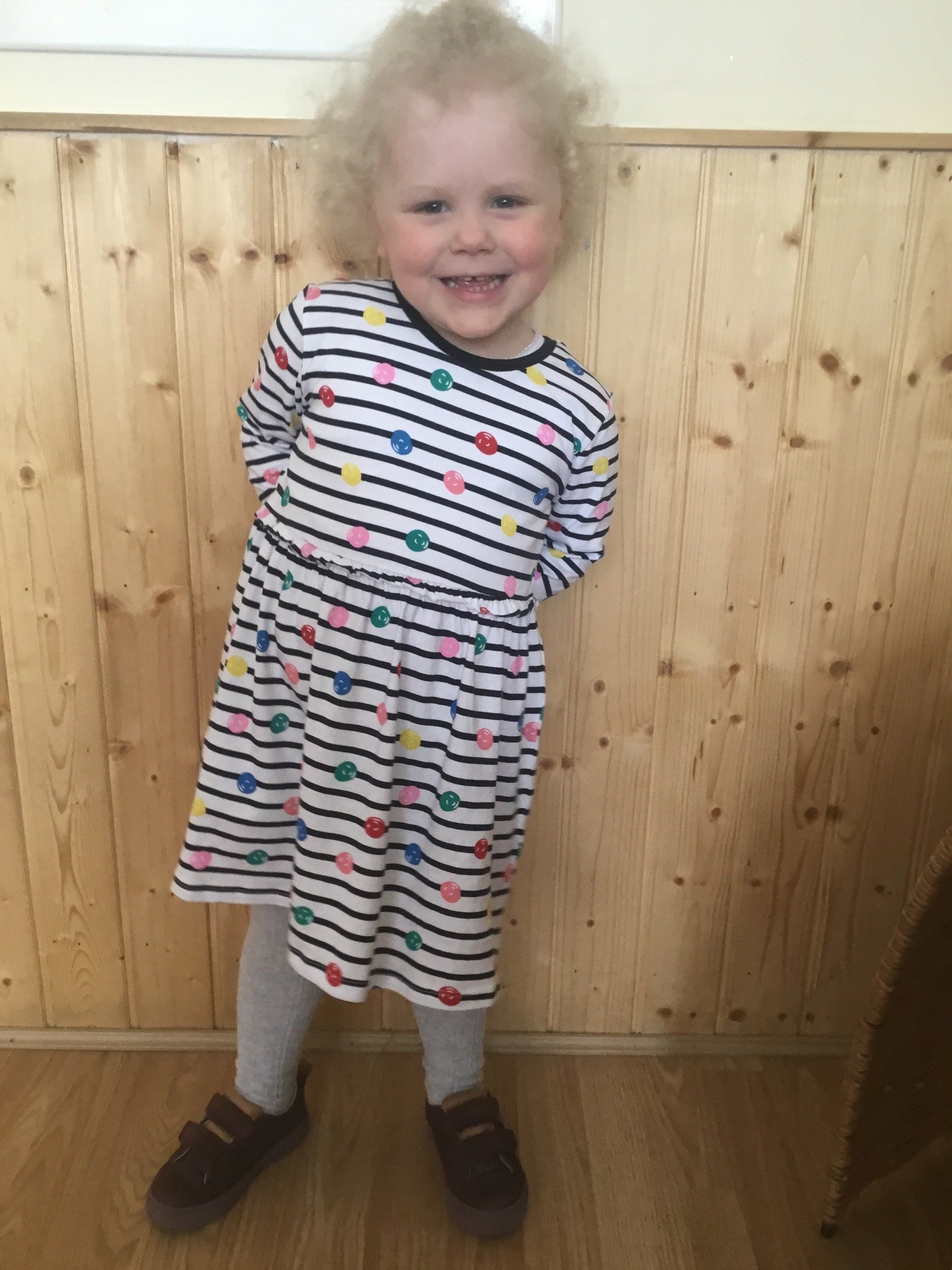 Smiling child in a striped dress standing against a wood-paneled wall