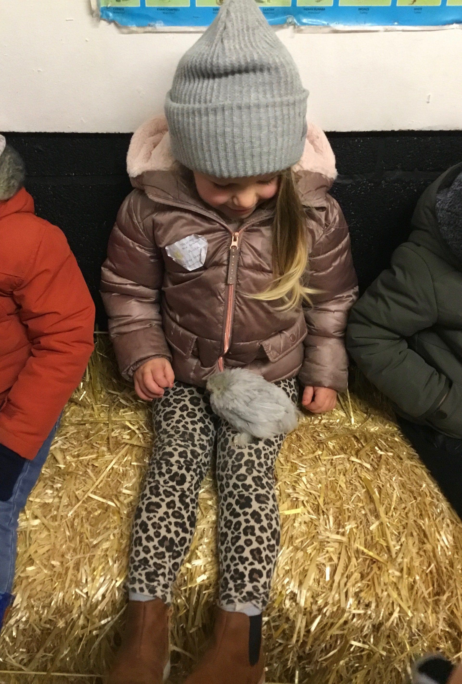 Child in gray beanie and brown jacket sitting on a hay bale, looking down at leopard-print leggings.