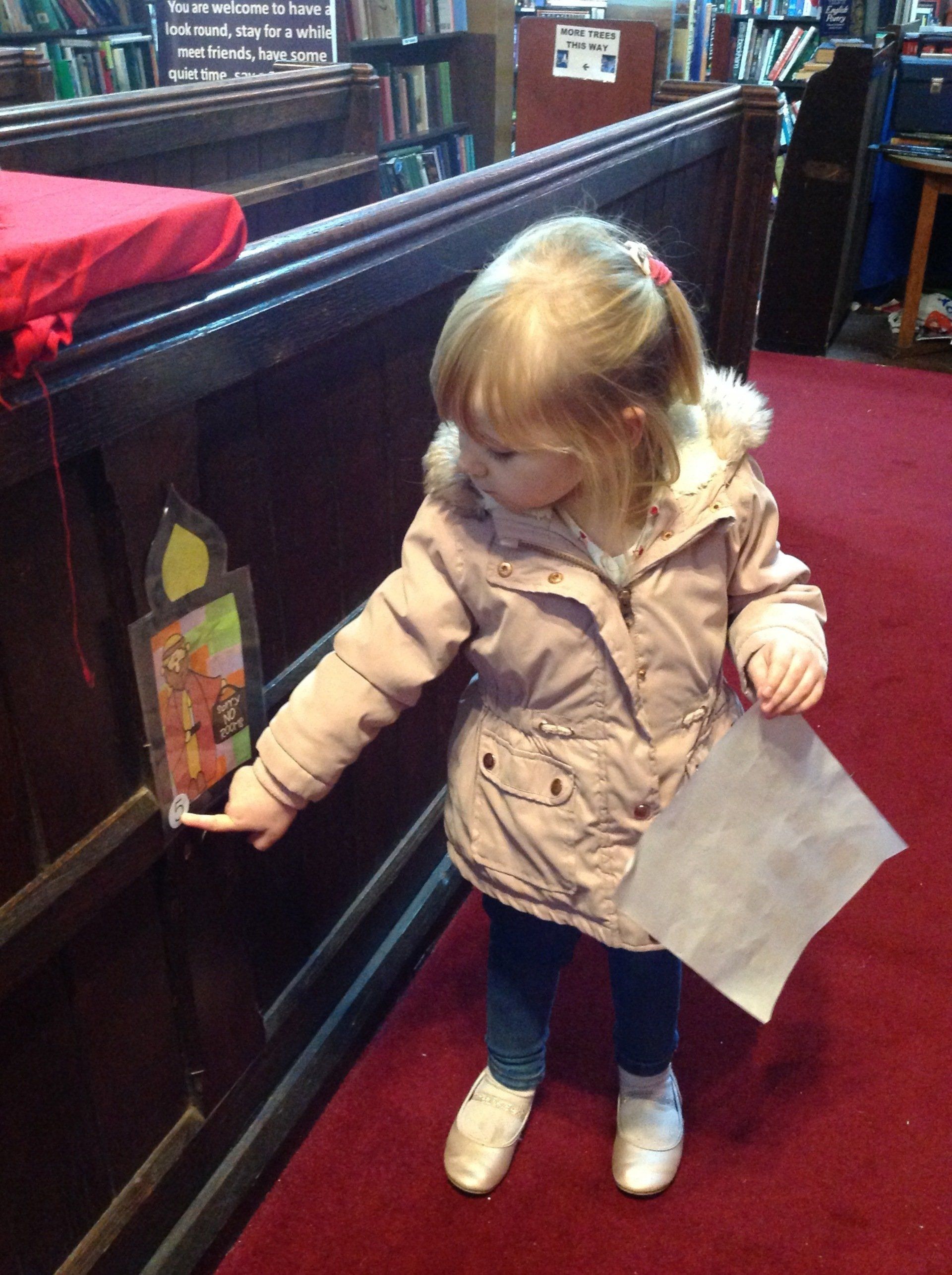 Young child in a tan coat reaching toward a bottle display in a shop, holding a clear bag