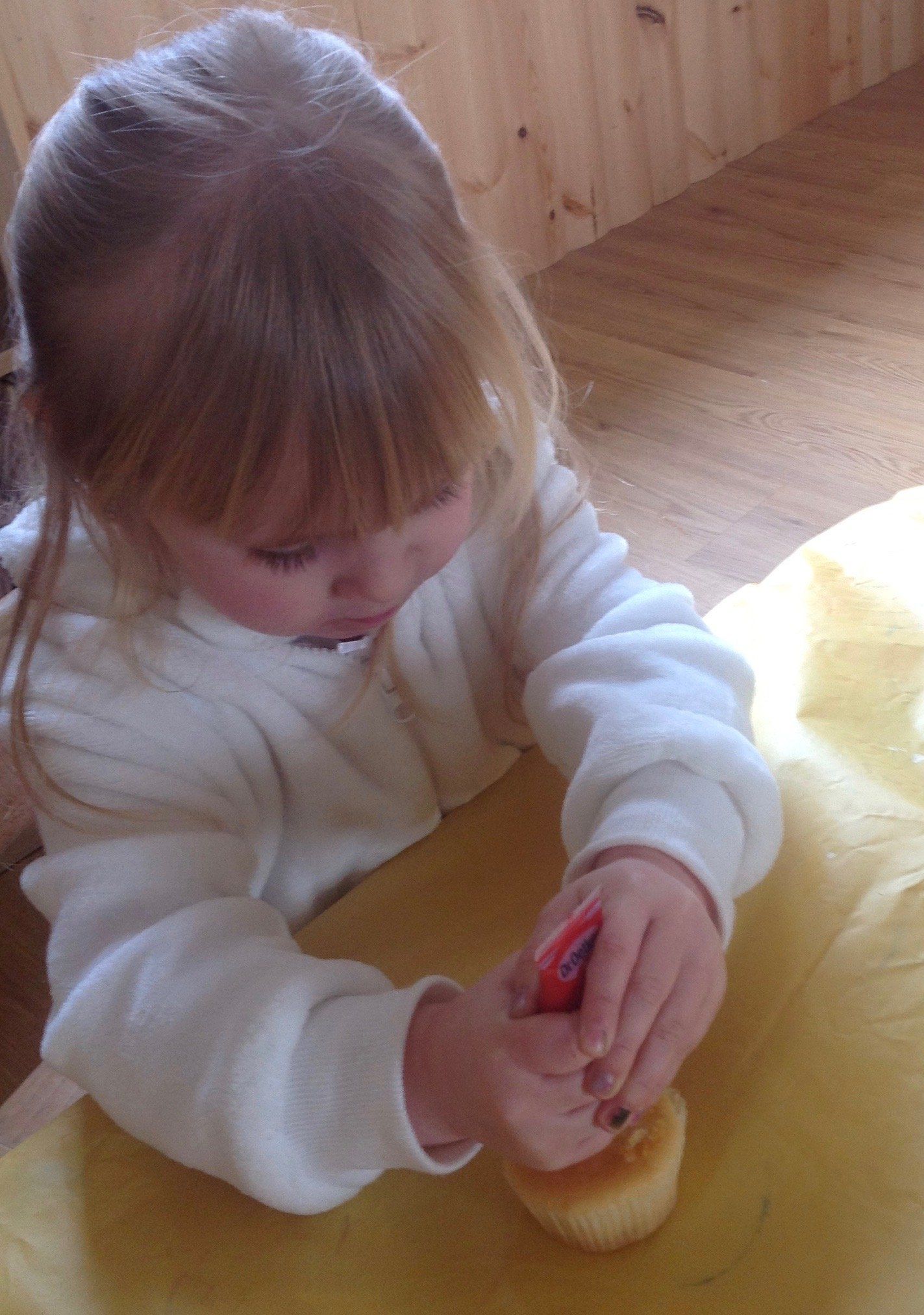 Toddler in a white sweater using a red tool on a cupcake on a wooden floor