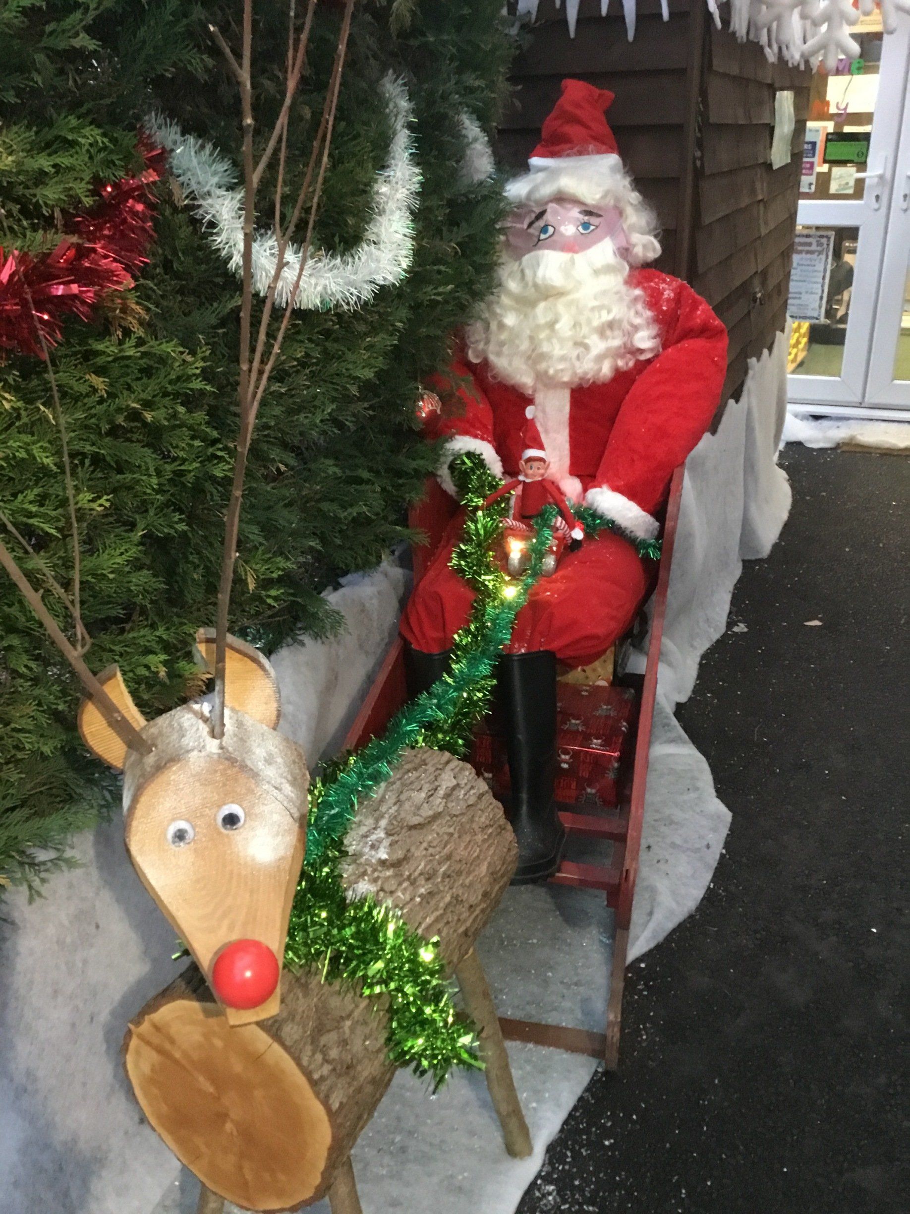 Santa in red suit seated beside a decorated reindeer display on a sidewalk