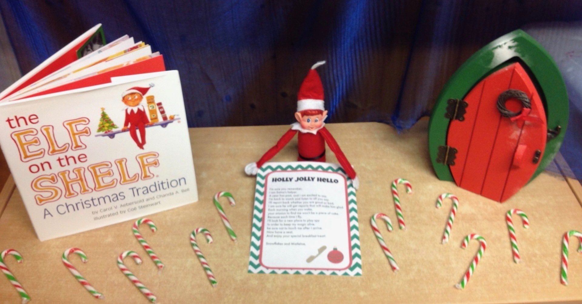 Elf on the Shelf display with candy canes, a red elf, book, note, and a green door on a wooden table