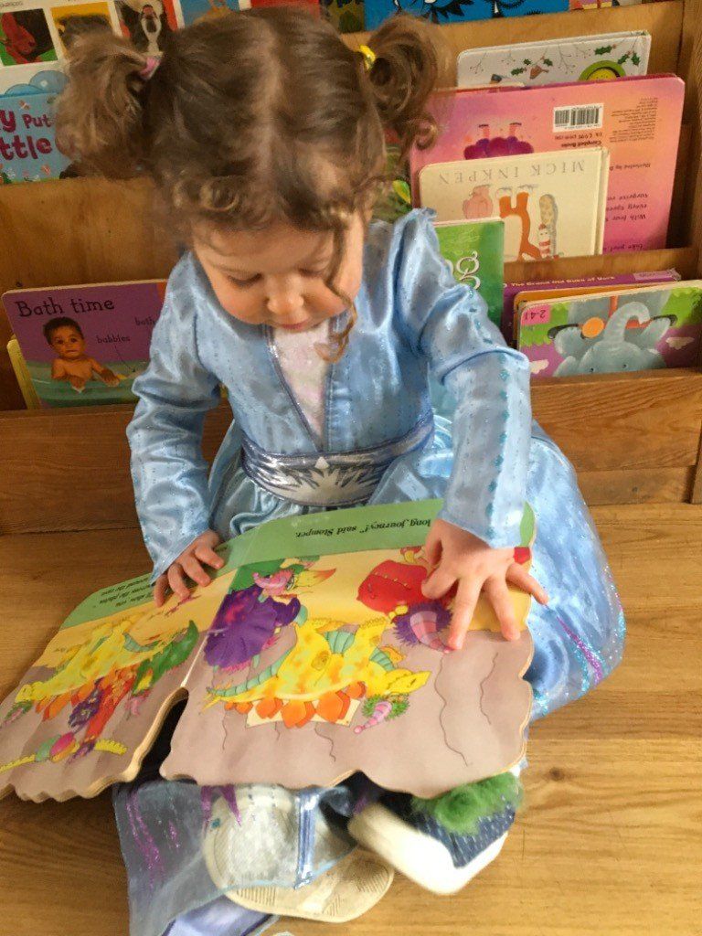 Child in a blue dress reading a colorful pop-up book, surrounded by books on a shelf.