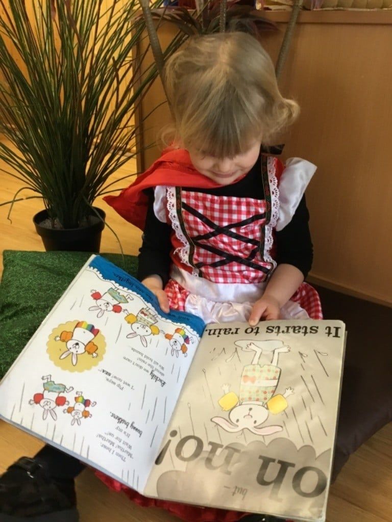 Child in a red costume reading an open picture book beside a potted plant.
