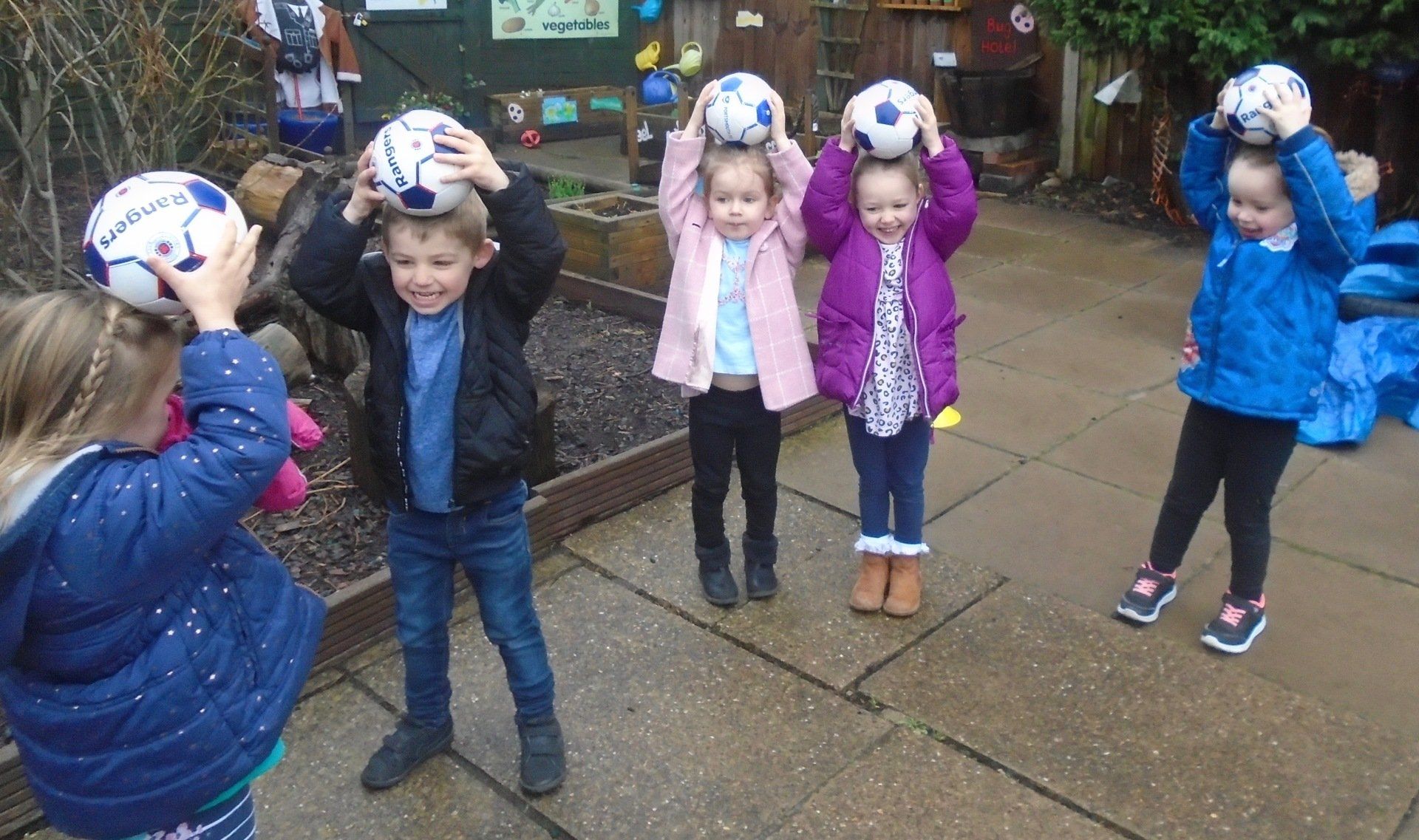 Children outdoors balancing soccer balls on their heads in a line on a wet playground.