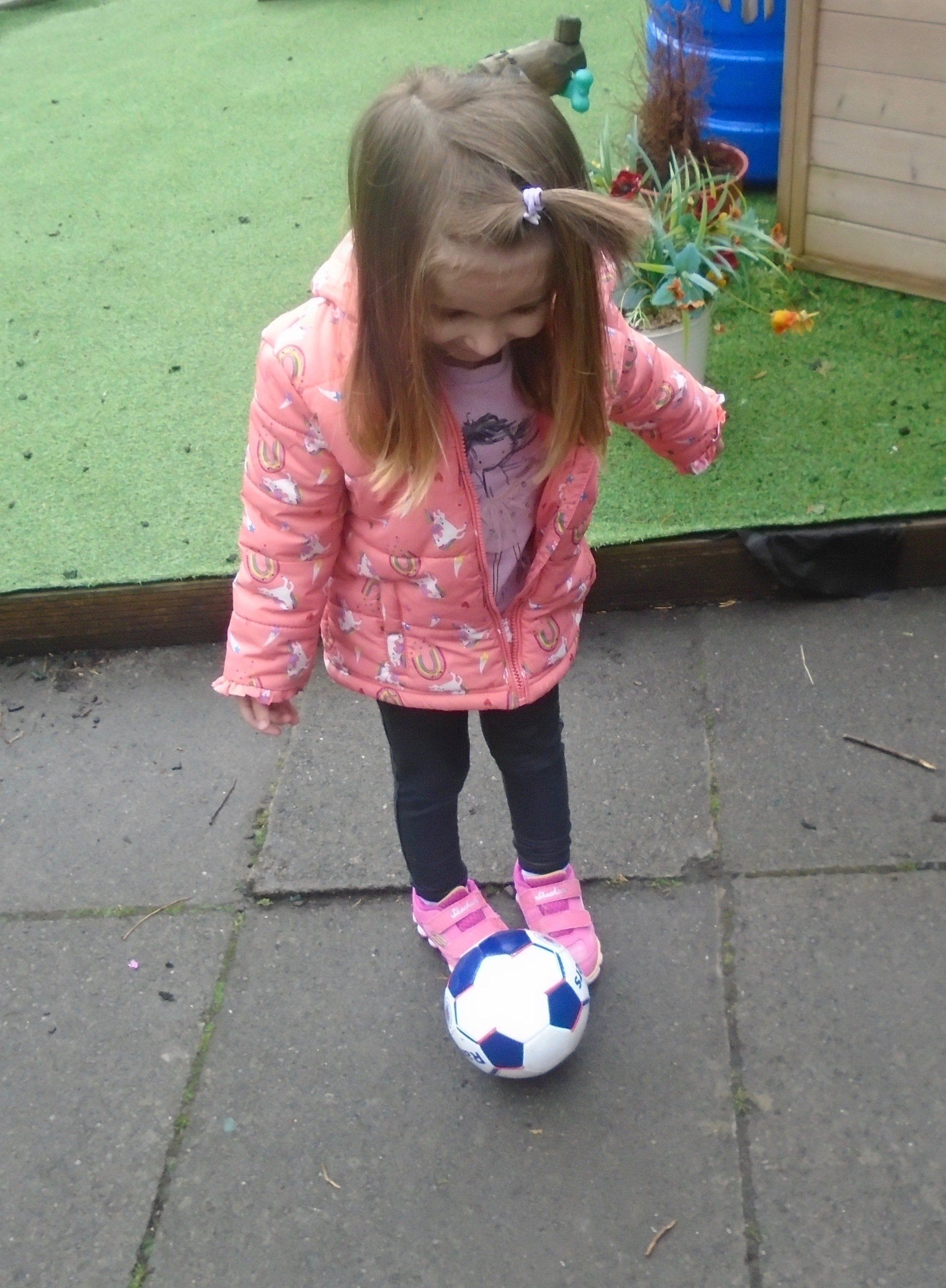 Child standing on a soccer ball outdoors on a sidewalk, wearing a pink jacket and black leggings.