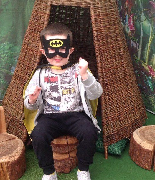 Child in a Batman mask sits on a woven chair, holding small objects in a playful setting.