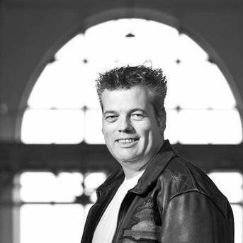 Een glimlachend persoon in een leren jas staat voor een helder verlicht boogvormig raam.