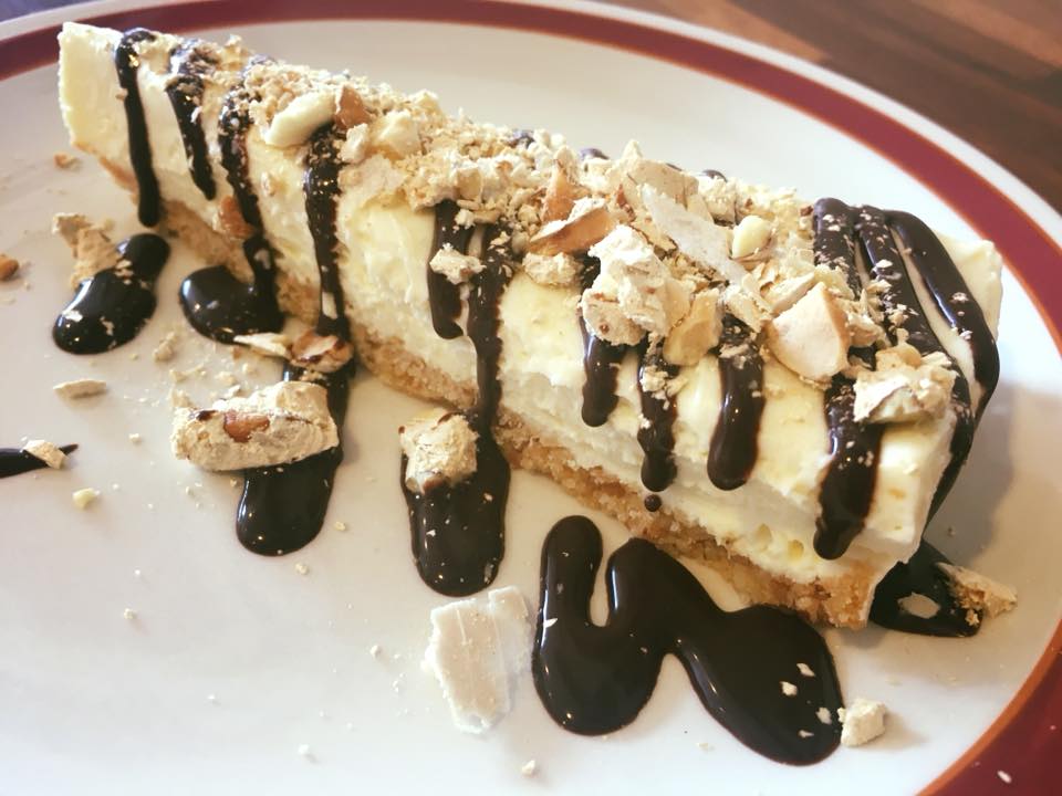 Cheesecake slice with graham cracker crust, drizzled with chocolate and topped with chopped nuts.