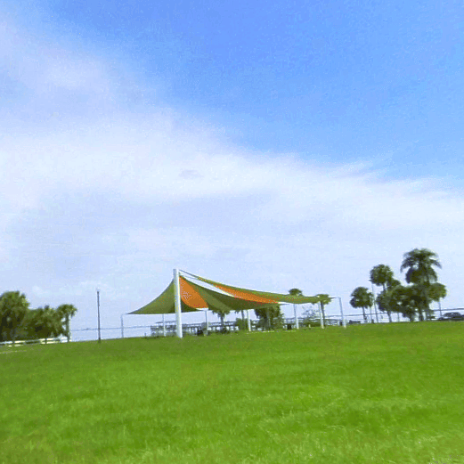 Green grassy park with a shade structure, palm trees, and a blue sky with clouds.