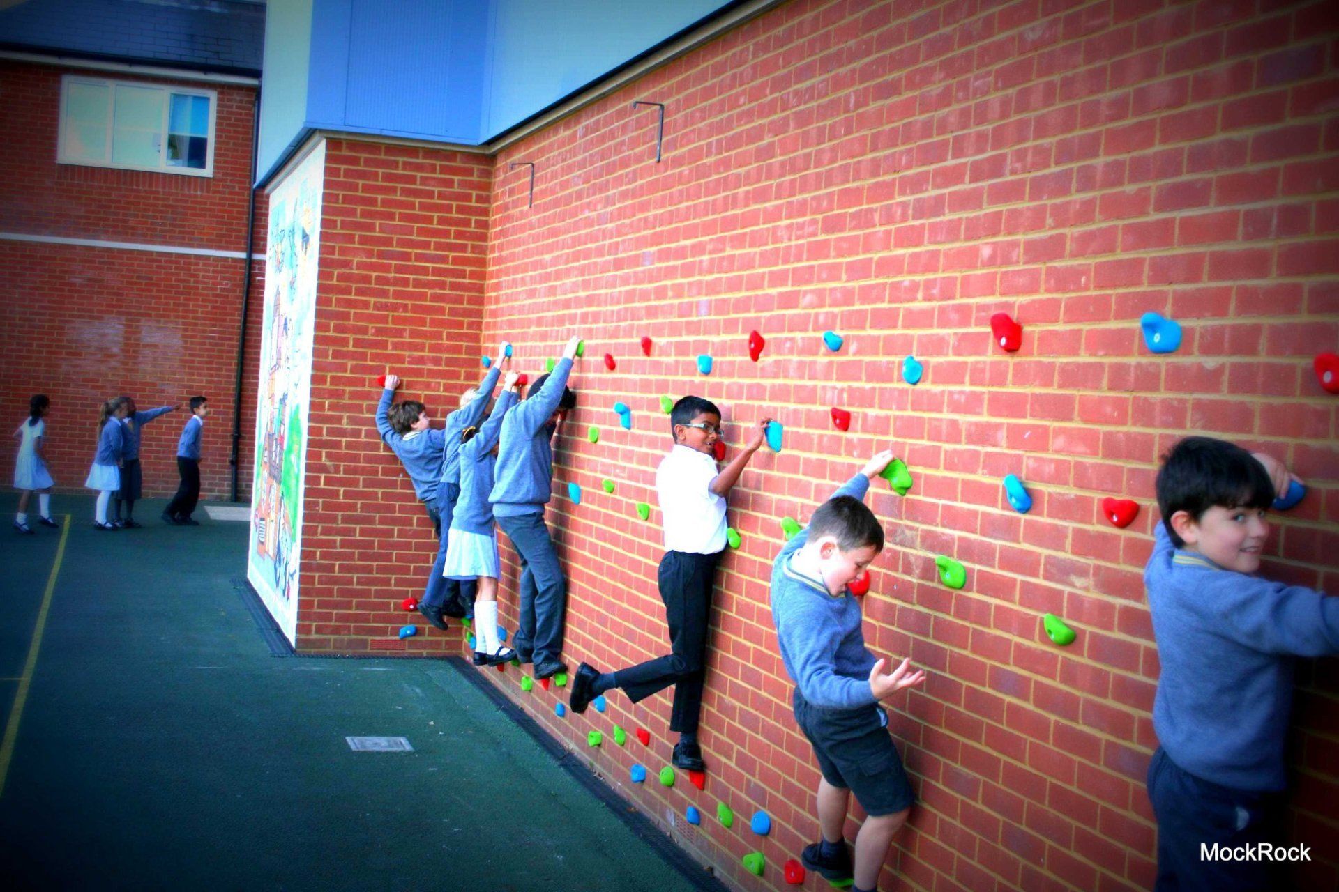 Children climbing a brick wall with colorful climbing holds at an outdoor school.