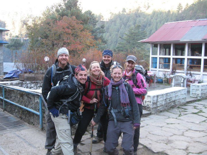 Group of hikers, smiling, posing together outdoors near a building with red roof, stone structure in mountainous region.