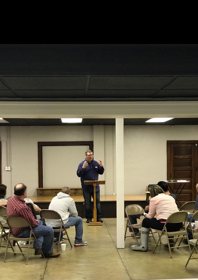 A man is standing at a podium talking to a group of people sitting in chairs.