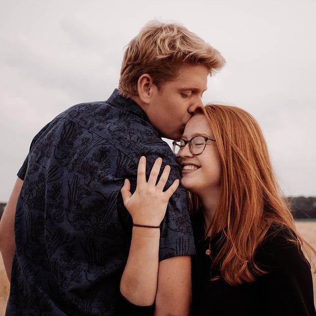 Een man kust een vrouw op haar voorhoofd; beiden glimlachen. Ze bevinden zich in de buitenlucht, met een veld op de achtergrond.