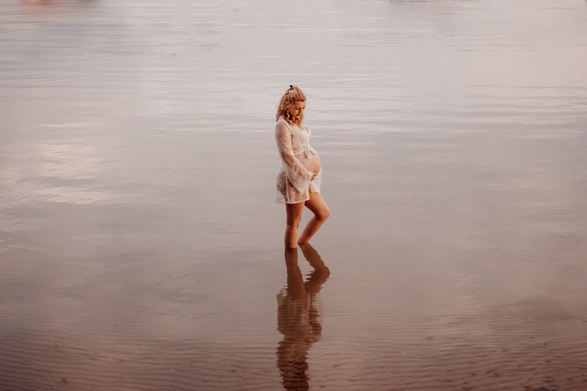 Een zwangere vrouw staat in ondiep water en draagt ​​een witte jurk. Zachtroze licht, de reflectie is zichtbaar.