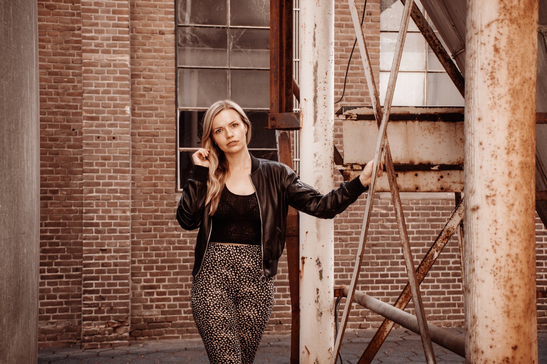 Vrouw in leren jas en broek met patroon poseert naast roestige metalen constructies, met een bakstenen muur op de achtergrond.
