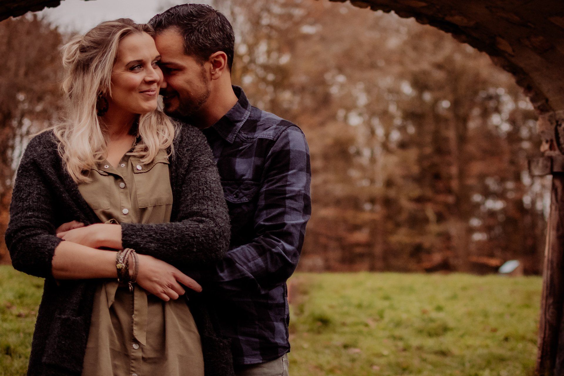 Een stel omhelst elkaar onder een stenen boog in een buitenomgeving. De vrouw glimlacht, de man kijkt naar beneden.