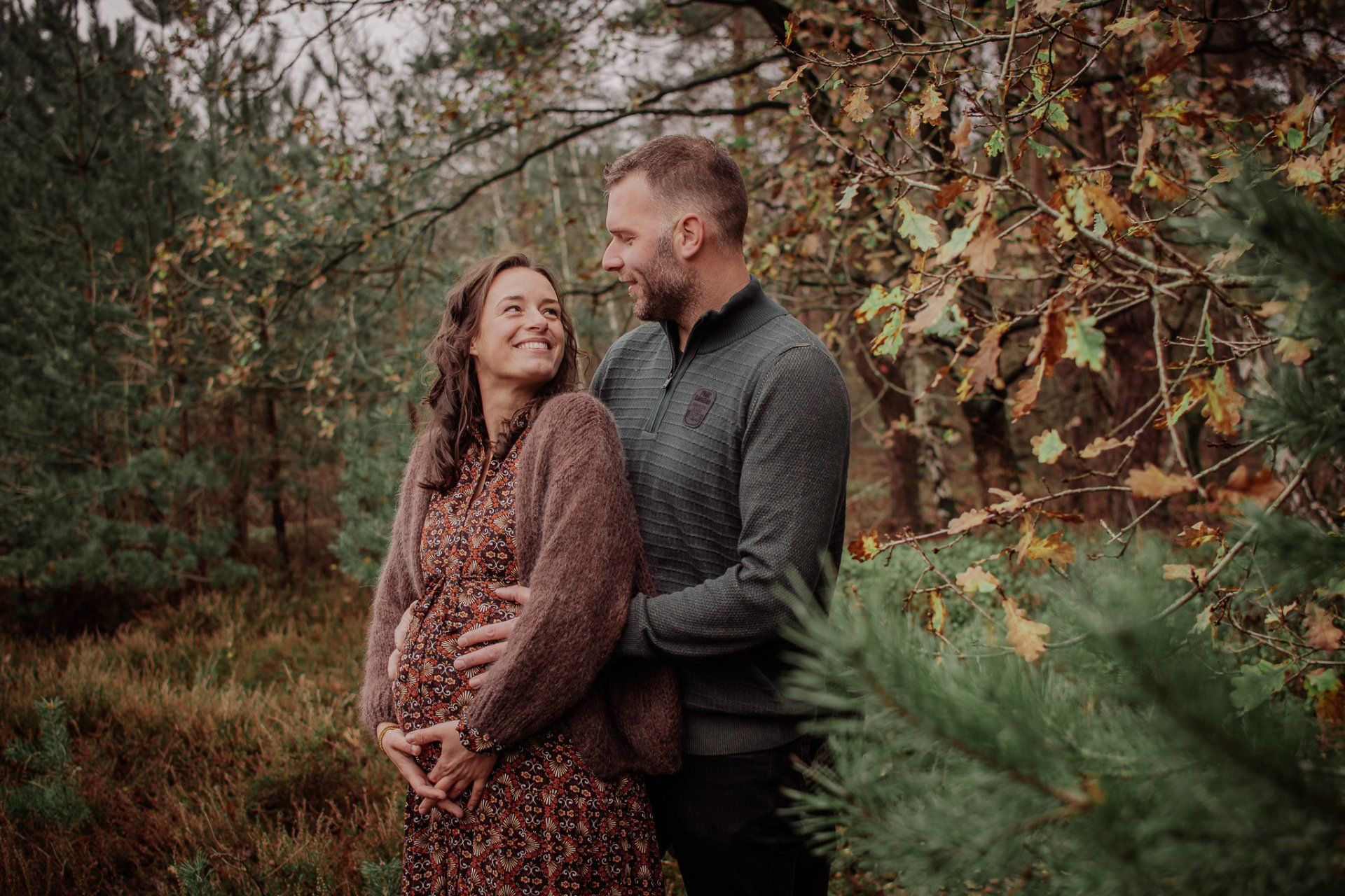 Een stel in een bos, de vrouw is zwanger, de man heeft zijn armen om haar heen geslagen, beiden glimlachen.