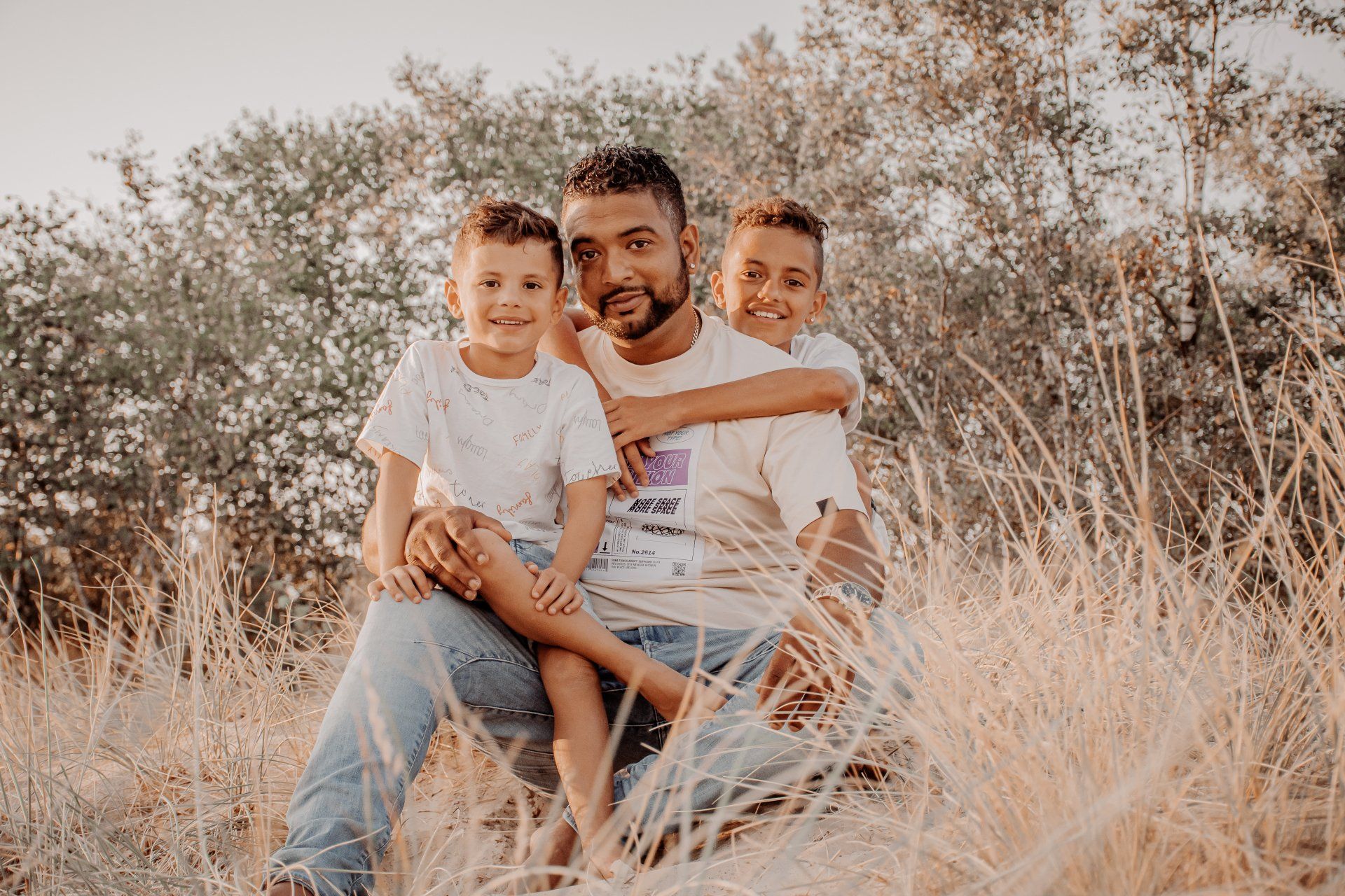 Een man zit met twee kinderen in een veld; ze lachen allemaal. De man omhelst de kinderen. Zacht, natuurlijk licht.