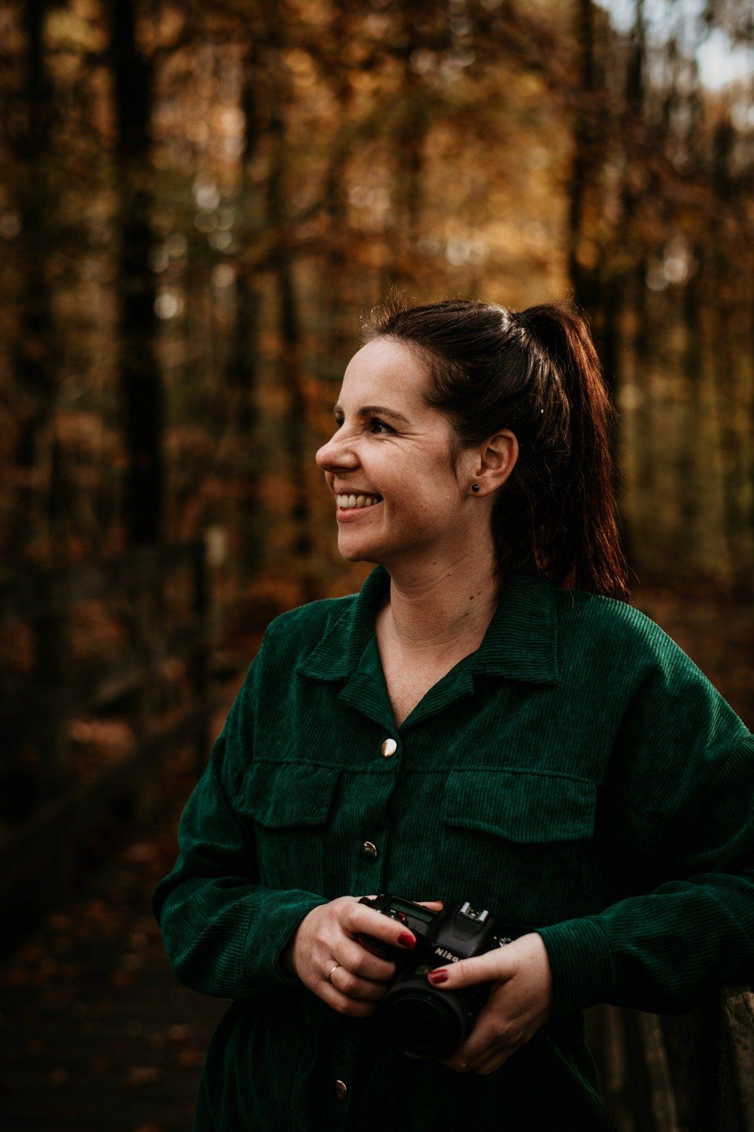Vrouw in een groene corduroy blouse, met camera in de hand, lacht in een herfstbos.