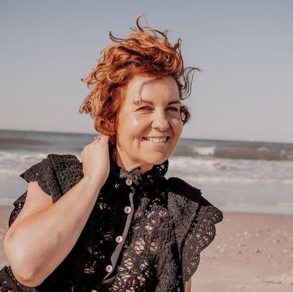 Vrouw met rood haar lacht op het strand, draagt ​​een zwart kanten topje, heeft haar hand in haar nek en de oceaan is op de achtergrond te zien.