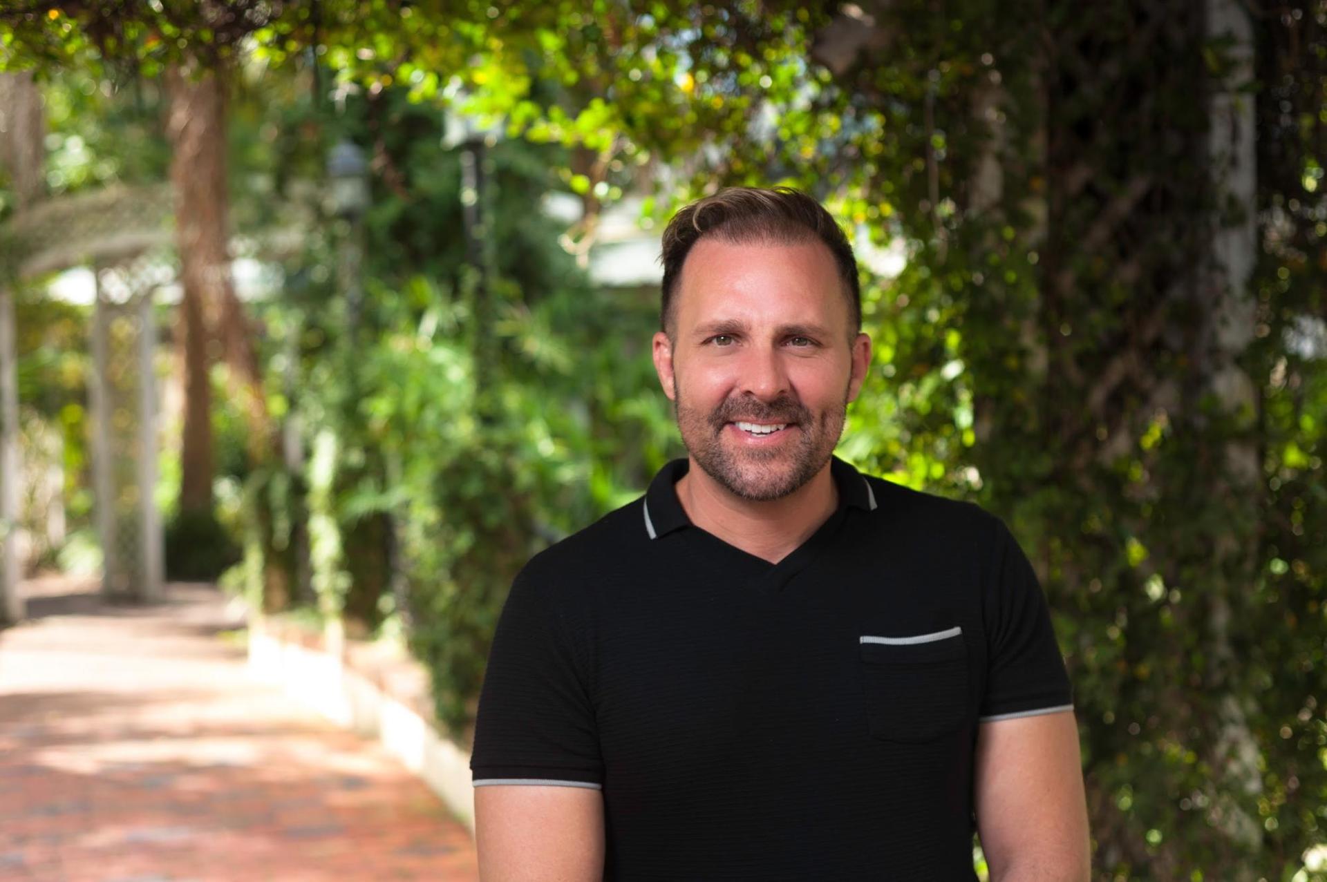 A man in a black shirt is standing in front of trees.