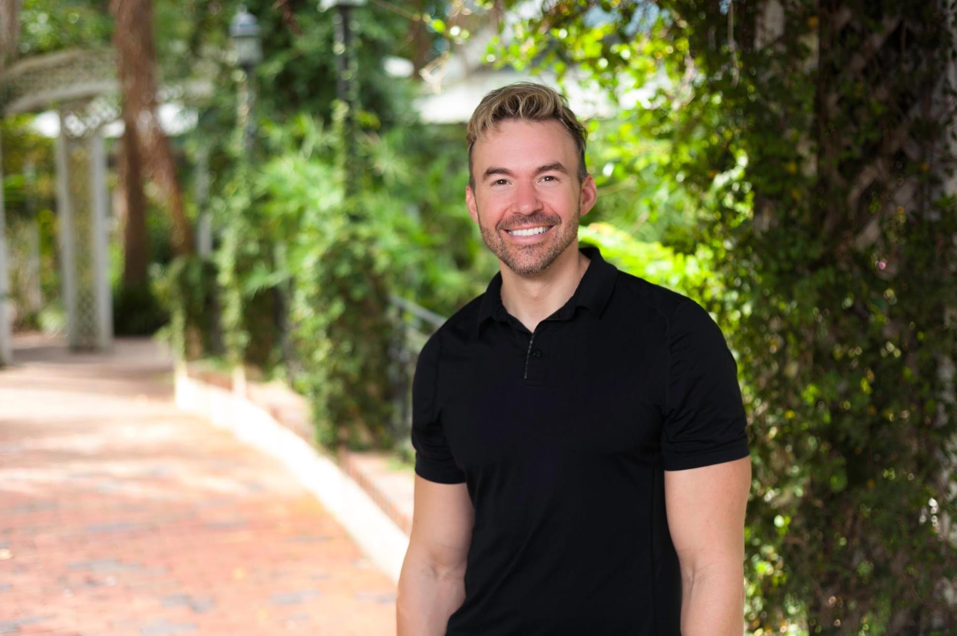 A man in a black shirt is standing on a sidewalk in front of trees.