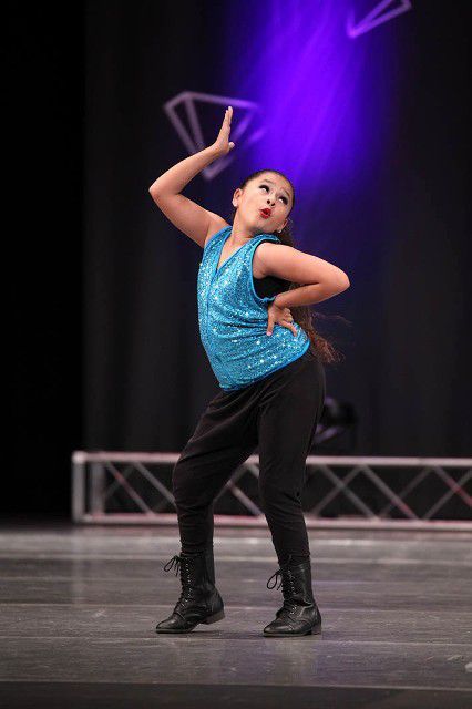 A young girl in a blue top and black pants is dancing on a stage.