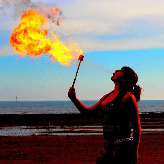 Fire performer blowing flames outdoors with the sea in the background, demonstrating fire entertainment acts available for UK events.