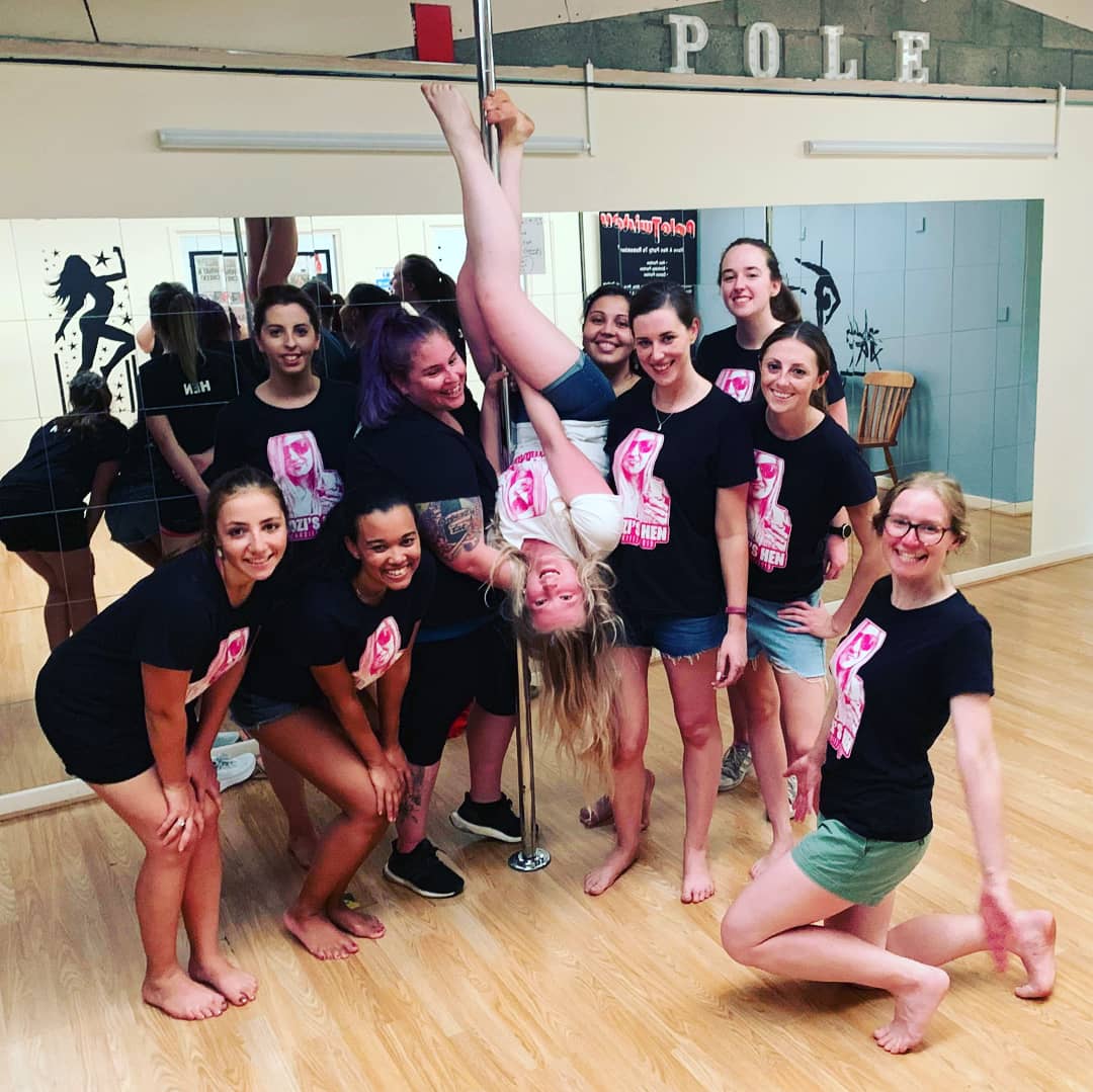 Group of friends enjoying a pole dancing party in a Cardiff studio, supporting a participant upside down on a pole and smiling together