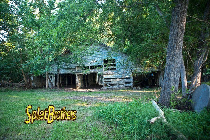 An old barn is sitting in the middle of a grassy field surrounded by trees.