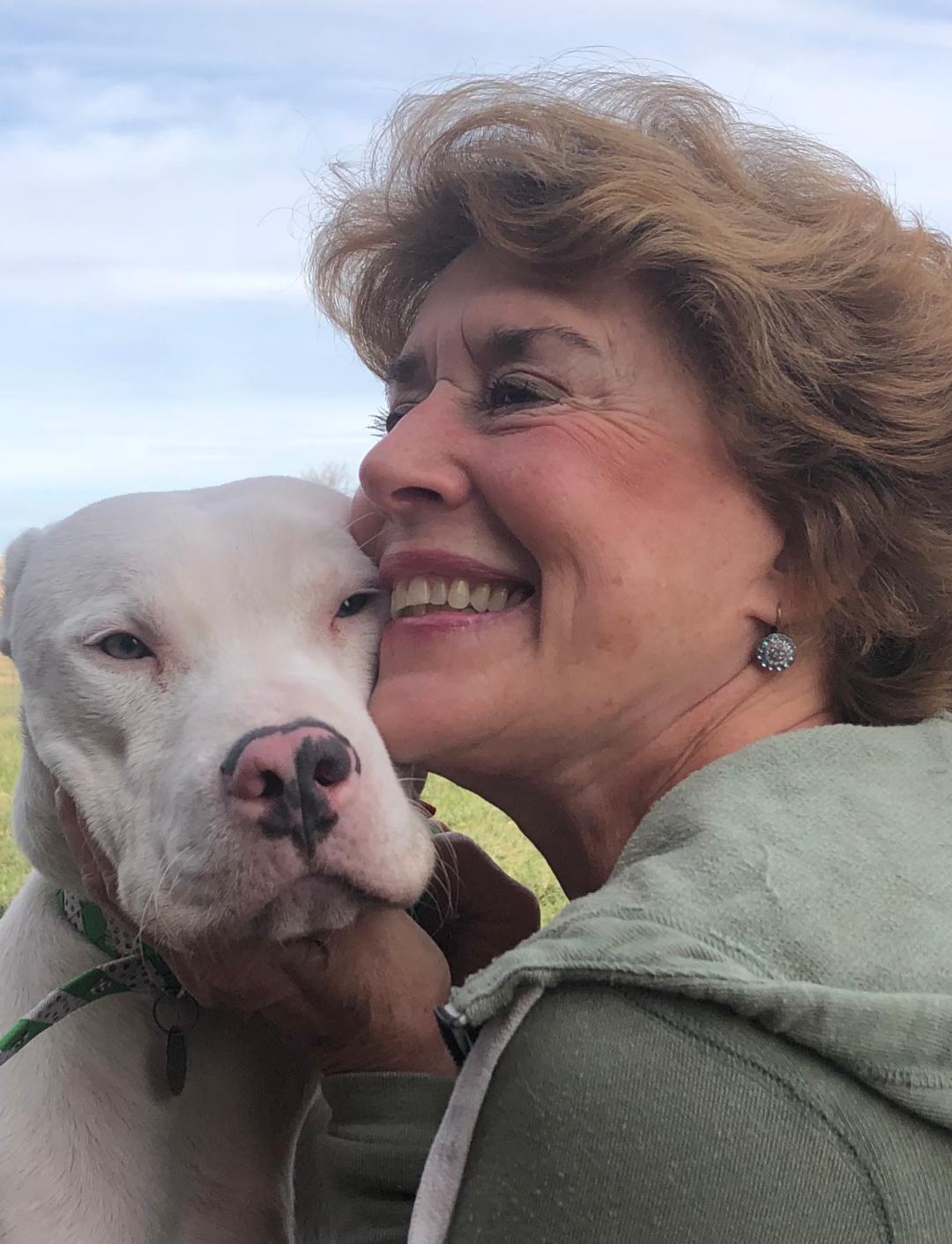 Woman with curly brown hair smiles while embracing a white dog outside.