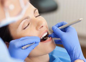 Woman in dentist chair, mouth open, dental tools present, blue gloves visible.