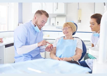 Dentist showing dentures to older woman in dental chair, assistant present. Bright, clean dental office.
