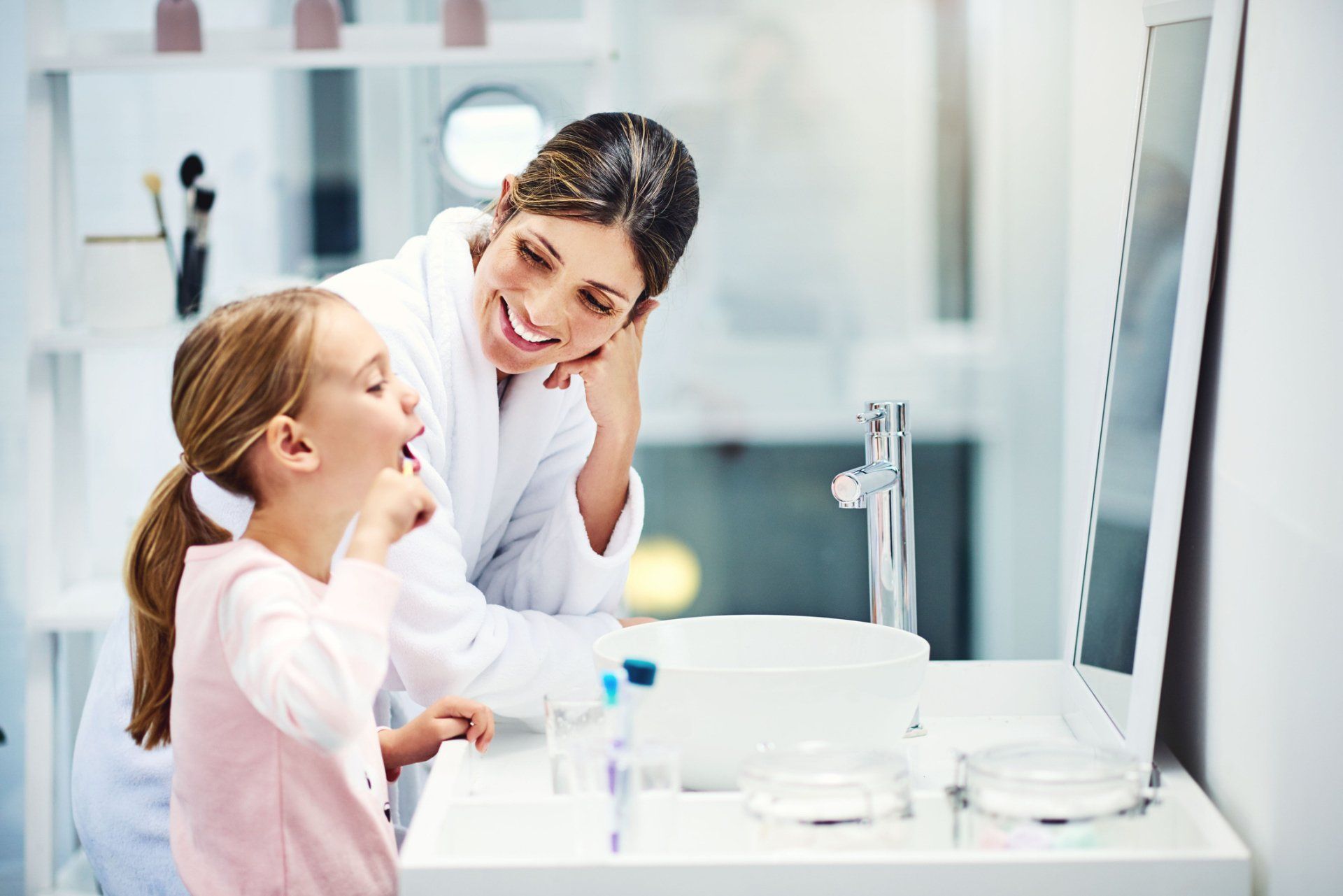 Woman watches a young girl brush her teeth in the bathroom, smiling, near a mirror.