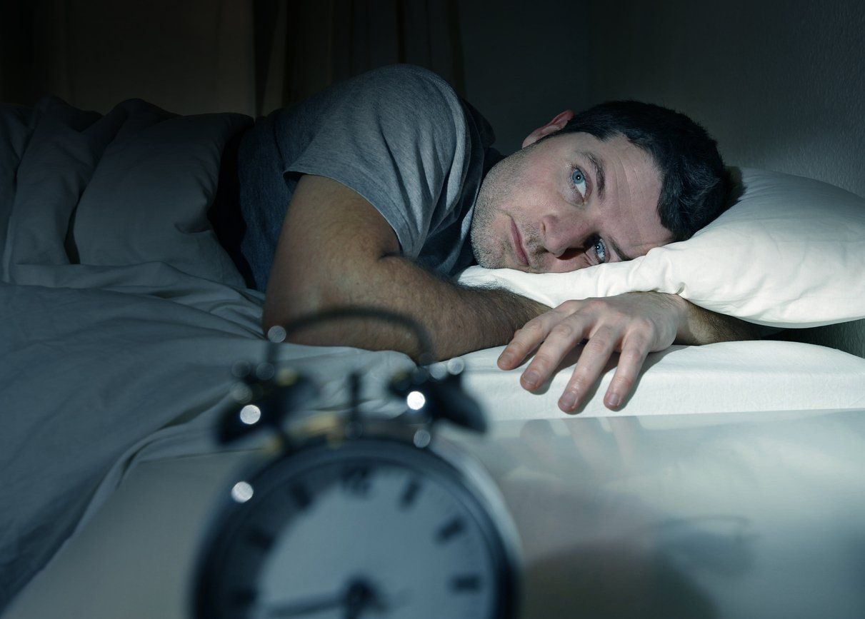 Man lying awake in bed, looking at alarm clock; concerned expression.