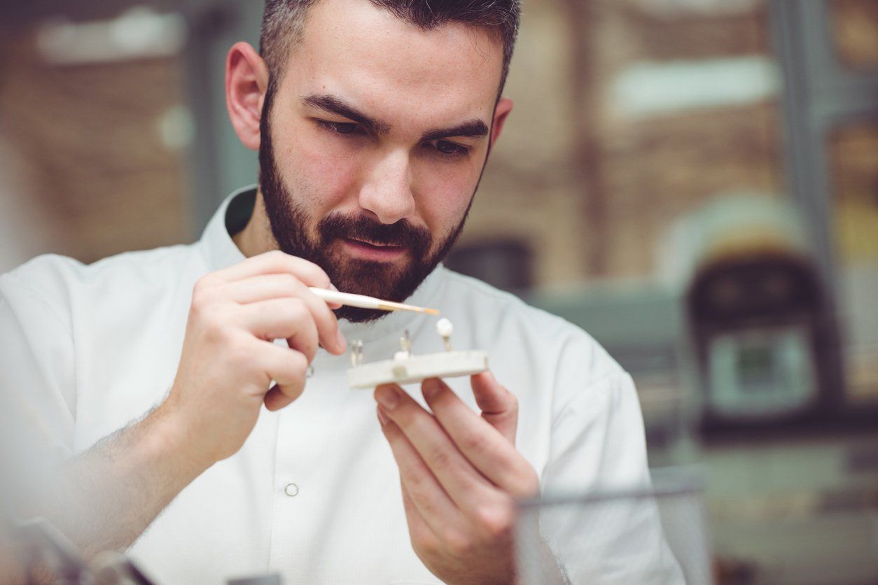 Man in white coat carefully working with small objects on a wooden base.