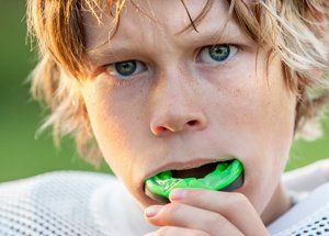Boy with blonde hair and green mouthguard looks intensely at the viewer, in a green field.