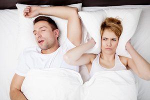 Man snoring in bed; woman covers ears with pillow, annoyed.