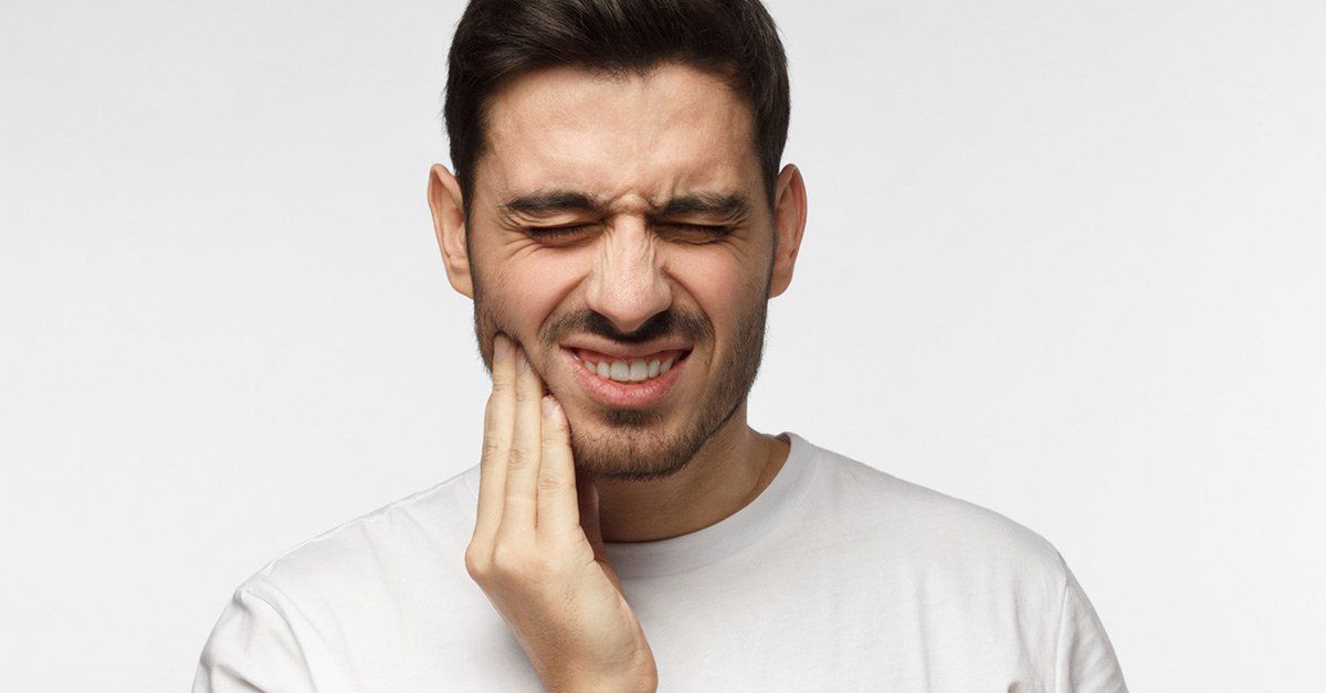 Man with pained expression, holding his jaw. White t-shirt, neutral background.