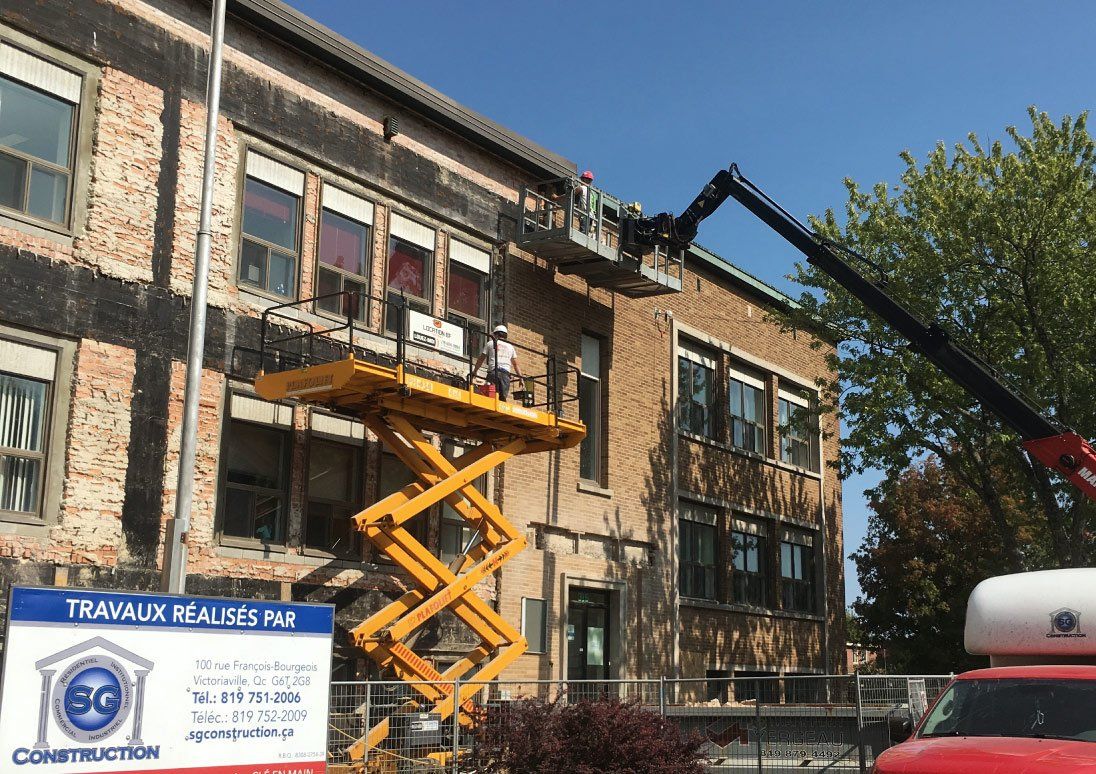 Des ouvriers du bâtiment sur une nacelle, réparant la façade en briques d'un bâtiment scolaire.