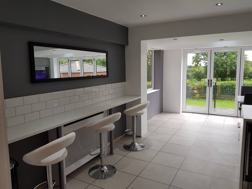 A long breakfast bar along a wall with modern white tile splashback, and then mid-grey painted walls. At the breakfast bar stands three modern, tall stools.