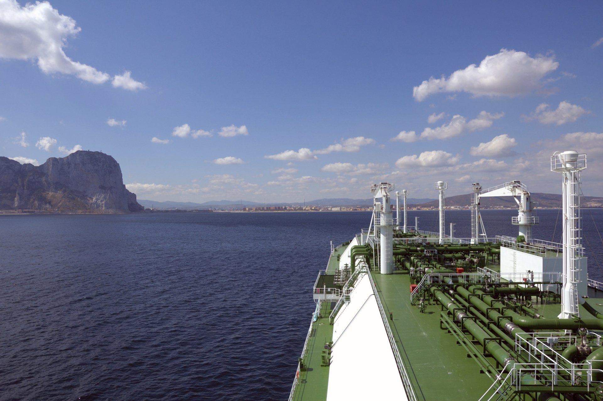 LNG tanker sailing on blue water towards a rocky coastline under a partly cloudy sky.