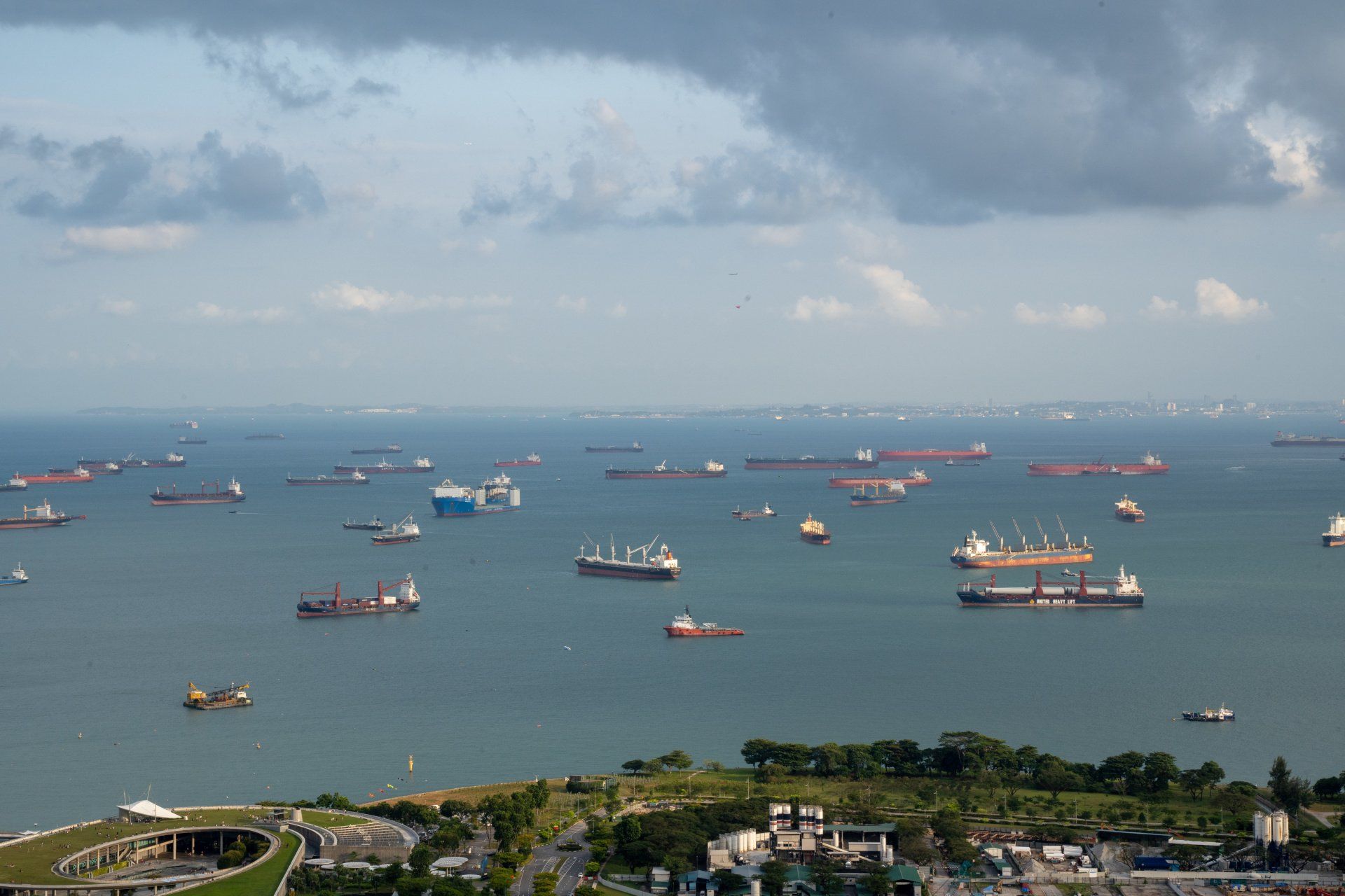 Shipping vessels scattered across a blue ocean, near a coastline under a cloudy sky.